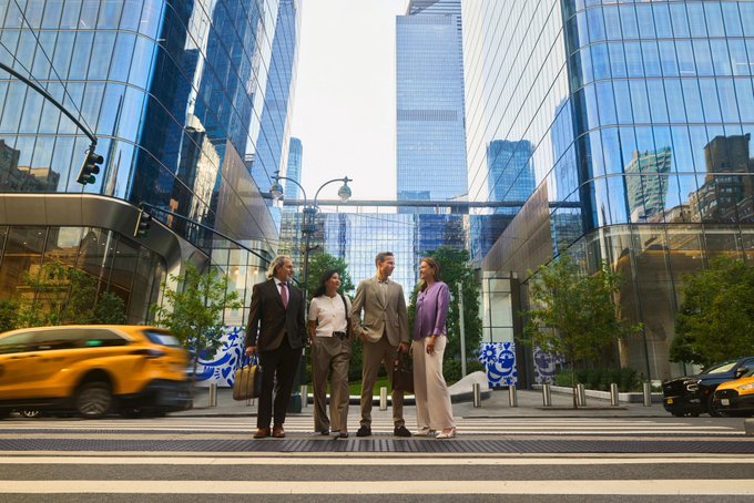 Four well-dressed business professionals standing together on a city crosswalk in front of modern glass skyscrapers, with a yellow taxi driving by.