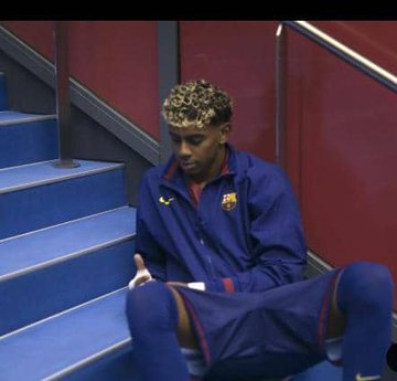 A man with curly bleached hair, wearing a blue and red Barcelona tracksuit, sits on blue stadium steps.