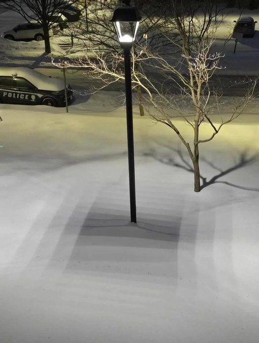 Nighttime aerial view of a snow-covered street and parking lot with an illuminated lamppost, bare tree, and a parked police vehicle visible on the left.