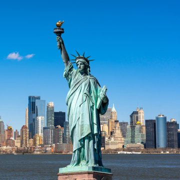 The Statue of Liberty standing prominently in the foreground with the New York City skyline visible behind it under a clear blue sky.