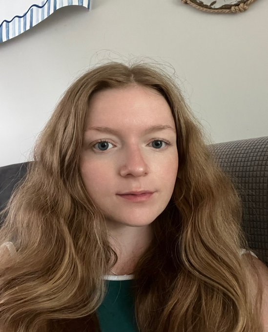 A young woman with long reddish-blonde hair looks directly at the camera with a calm, neutral expression, sitting on a gray couch. The casual selfie aligns with the post's self-deprecating humor about having a small, dedicated audience of "10 people" greeting her in the morning.