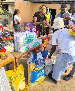 People gather around tables stacked with household goods—including packs of toilet paper, pet food labeled "Pet Lover," and a yellow "Seafood" bag—as a woman hands out items to recipients during what the post describes as an outreach to feed orphans.