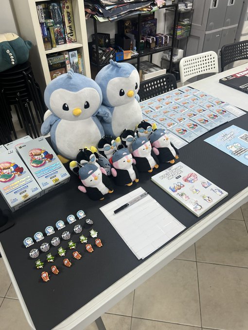 A table display featuring large and small blue penguin plush toys, video game boxes, trading cards, small character badges, a guestbook, and a marker, likely at a fan event.