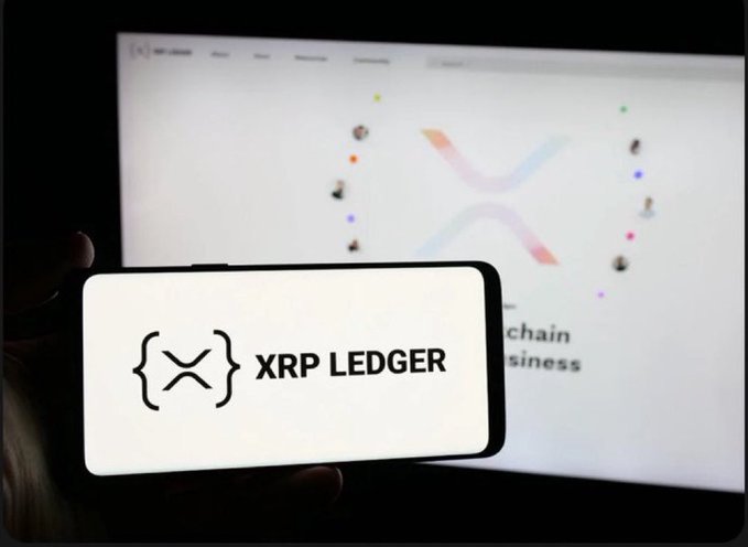 A hand holding a smartphone displaying the "XRP LEDGER" logo in front of a computer screen showing the XRP website.