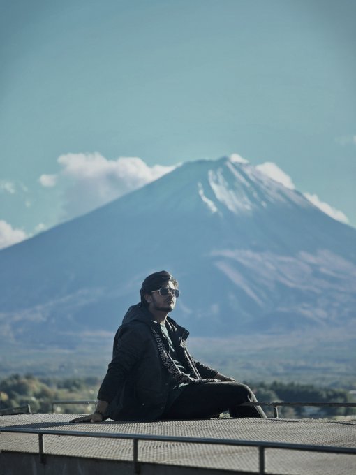 A man wearing sunglasses and a dark jacket sits on a rooftop platform with a large snow-capped mountain looming in the background under a blue sky.