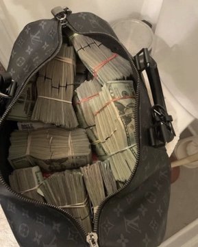 An open Louis Vuitton monogram duffel bag overflowing with numerous rubber-banded stacks of U.S. dollar bills.