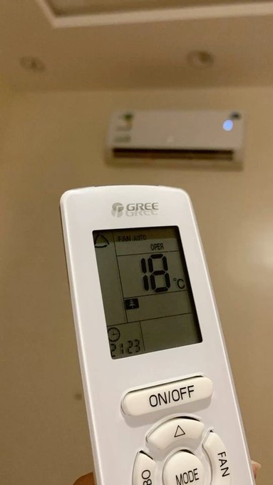 A close-up of a hand holding a GREE air conditioner remote control showing 18°C, with the wall-mounted AC unit visible in the background.