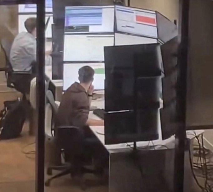 A person sitting in an office cubicle surrounded by multiple computer monitors displaying various screens.