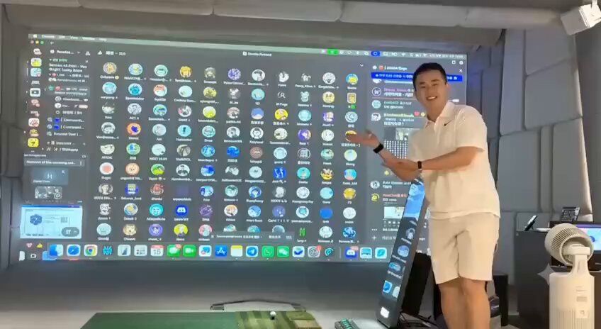 The image features a man standing next to a large screen displaying various computer icons and logos. The man appears to be giving a presentation or explaining something related to these icons, possibly about a new app or software. The screen behind him is black with white text on it, which could be part of the presentation or explanation he's providing.