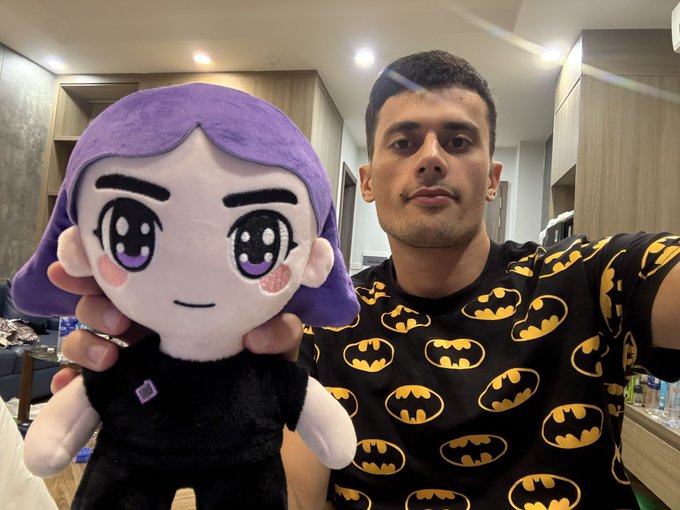 A man wearing a black t-shirt with yellow Batman logos is taking a selfie while holding up a plush doll with purple hair, large anime-style eyes, and a black outfit.