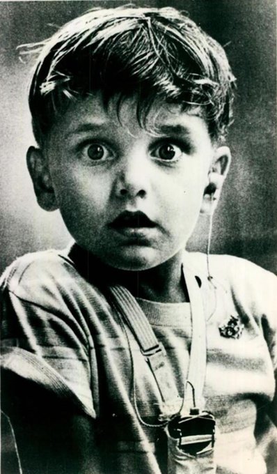 A black and white vintage photograph of a young boy with messy hair and a surprised expression, wearing overalls with a strap visible across his chest.
