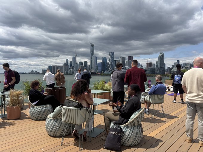 A rooftop gathering shows people relaxing on outdoor furniture with a view of the Toronto skyline, including the CN Tower, across the water under cloudy skies.