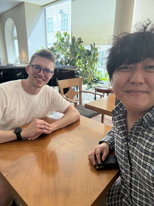 Two young men sitting at a wooden table in what appears to be a cafe or restaurant, with one wearing glasses and a white t-shirt, and the other in a plaid shirt.