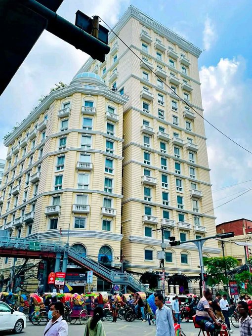 A street-level photograph of a large, ornate yellow multi-story building with classical architectural details, located in a busy area with people, rickshaws, and a pedestrian overpass visible at the base.