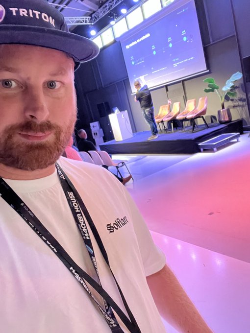 A man wearing a 'TRITON' hat and lanyards takes a selfie in front of a conference stage with a presentation screen and empty chairs in the background.
