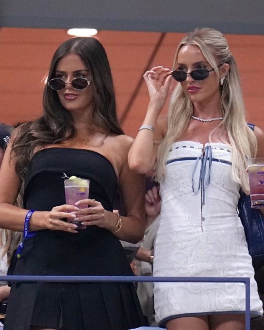 Two blonde women in fashionable outfits and sunglasses, holding drinks, seated in the stands at what appears to be a tennis match.