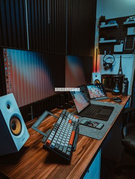 A desk setup features dual monitors displaying colorful screens, a mechanical keyboard, a laptop, studio monitors, and warm ambient lighting.
