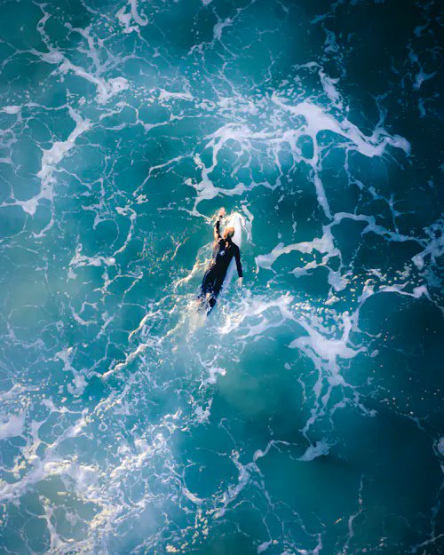 An aerial view shows a surfer paddling through foamy turquoise ocean waves, appearing tiny against the vast expanse of water.