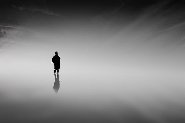 A black and white artistic image showing the silhouette of a person standing alone in a misty, ethereal void with their reflection visible below.