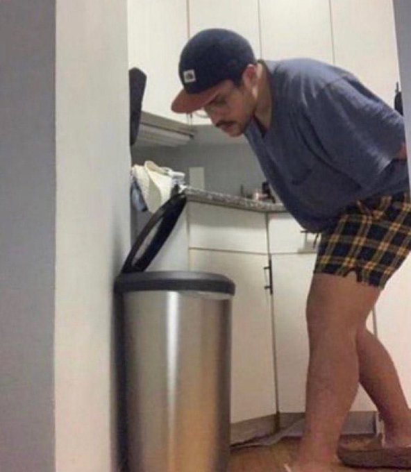 A man wearing a baseball cap, gray t-shirt, and plaid boxer shorts stands in a kitchen next to a stainless steel trash can, looking down at something on the floor.