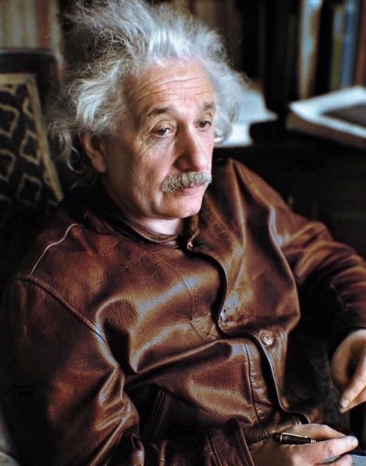 A colorized photograph shows an elderly Albert Einstein wearing a brown leather jacket, seated and holding a pen.