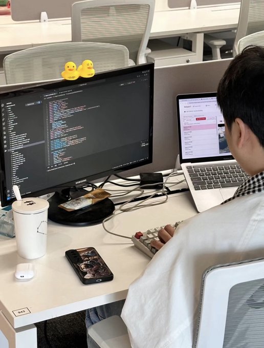 A person working at a desk in an office cubicle, viewing code on a Samsung monitor with rubber ducks on top, a laptop beside it, a phone, AirPods case, and a drink cup nearby.
