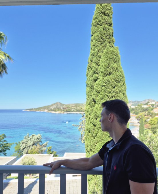 A young man in a dark polo shirt stands on a balcony overlooking a stunning blue sea with a coastline and tall cypress tree visible in the background.