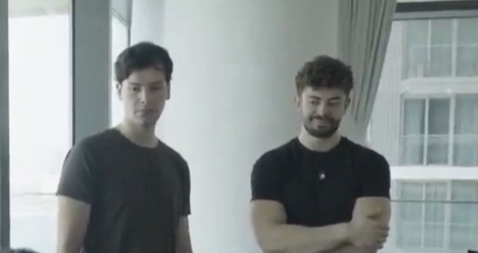 Two young men, both wearing dark t-shirts, stand casually in a bright indoor space with large windows in the background. The image likely represents the crypto traders or investors behind the claim of profiting 1,000 SOL after their portfolio reached 200k, suggesting a moment of quiet celebration or reflection on their trading milestone.