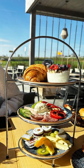A three-tiered serving stand on a sunny outdoor table holds a croissant, a parfait with berries, a plate with meats, cheeses, and tomatoes, and a bottom plate with assorted cheeses, fruits, and condiments.