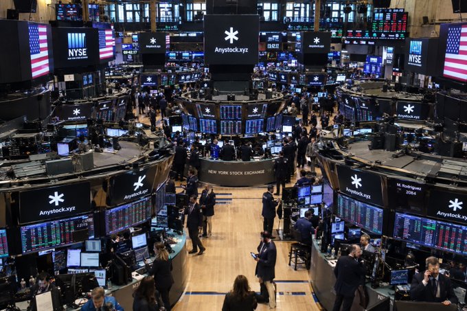 An overhead view of the New York Stock Exchange trading floor filled with traders, trading stations, and digital screens displaying NYSE and Anystock.xyz branding alongside American flags.