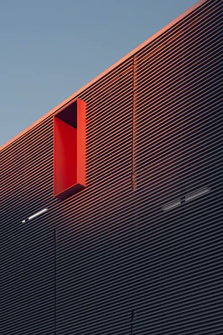 A minimalist architectural shot featuring a bright red rectangular vent protruding from a corrugated metal building facade against a clear sky.