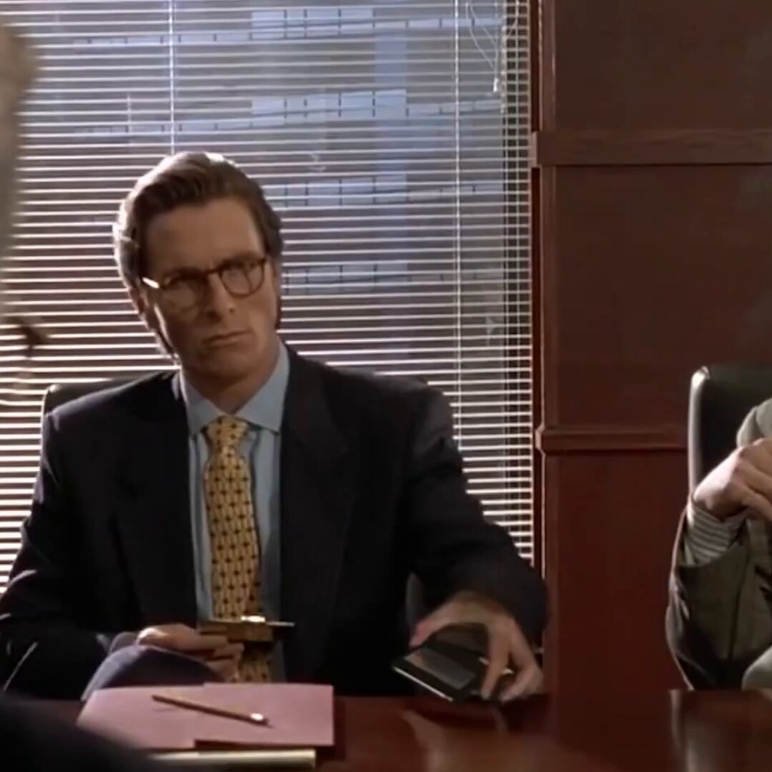 The image shows a man sitting at a desk with a book open in front of him. The man appears to be dressed in business attire, wearing glasses and a suit. There are two other people present in the scene - one person on the left side of the frame and another person on the right side. They seem to be engaged in conversation or observing something happening at the desk.