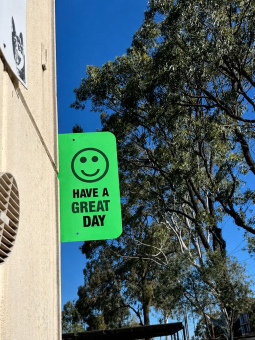 A bright green street sign featuring a smiley face and the text "HAVE A GREAT DAY" hangs on a beige building under a clear blue sky, perfectly matching the cheerful "gm!" (good morning) sentiment with its friendly greeting.