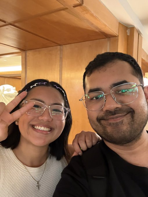 A selfie of a smiling young woman flashing a peace sign and a man with glasses, both posing indoors.