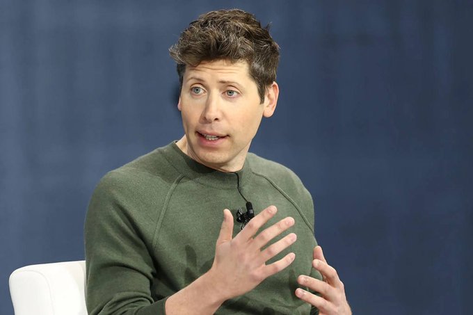 A man with curly brown hair, wearing a green sweater and a small lapel microphone, gestures with his hands while speaking against a dark blue backdrop. The image appears to depict Sam Altman, CEO of OpenAI, the company referenced in the post about the recent exodus of its ethics, safety systems, and mission alignment leaders.