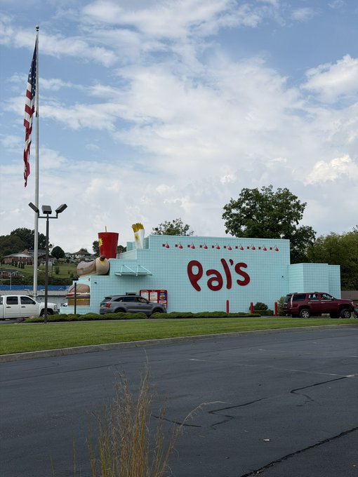 A light blue building with red 'Pal's' signage featuring a large hot dog and french fries statue on top, with an American flag flying nearby.