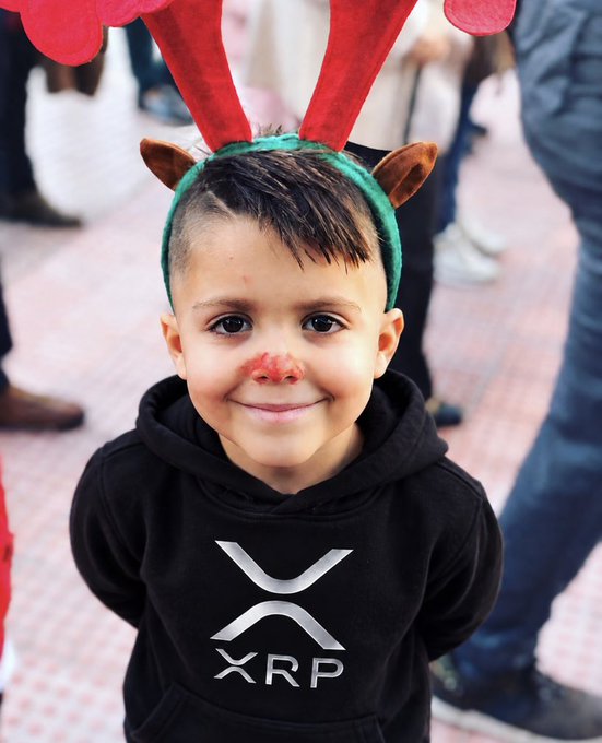 A smiling young boy wearing a black hoodie with the XRP logo and a festive reindeer antler headband with a red nose, celebrating his birthday with the XRP community. The image perfectly captures the post's sentiment, as Alan appears overjoyed while showcasing his XRP-themed attire.