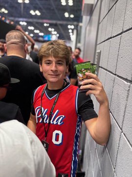 A young man wearing a red Philadelphia 76ers basketball jersey takes a selfie in what appears to be a crowded indoor arena or event venue.
