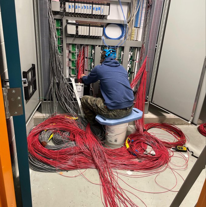 Photo of a technician sitting on a bucket while working on a complex telecommunications panel, surrounded by massive coils of red and gray cables.