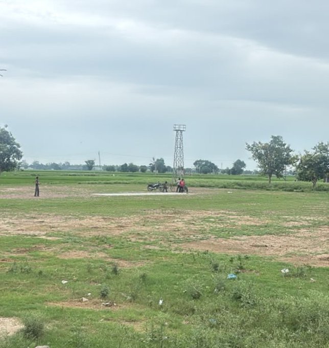 A wide landscape photo of a rural field with a few people gathered near a motorcycle under a tall metal transmission tower, with a bird flying in the cloudy sky to the left.