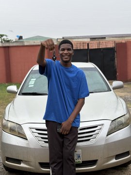 A young man in a blue t-shirt standing in front of a silver sedan, raising his fist in the air with a slight smile.