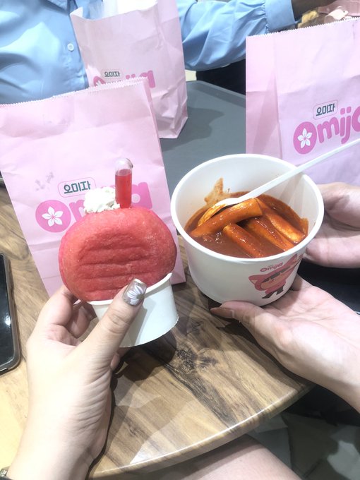 Two people sharing snacks at a table featuring 'omjo' branded pink paper bags, holding a pink dessert topped with a cherry in one hand and a cup of tteokbokki (spicy rice cakes) in the other.