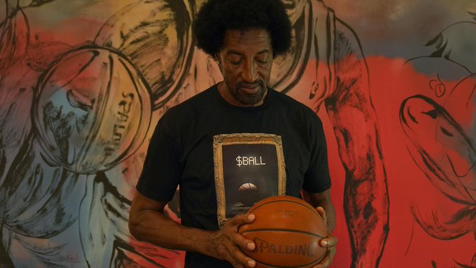 A man with an afro holding a Spalding basketball and wearing a black 'B$ALL' t-shirt in front of a red and black basketball-themed mural.