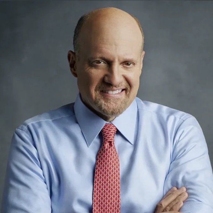 A smiling bald man in a blue dress shirt and red patterned tie poses for a professional headshot with his arms crossed against a gray background.