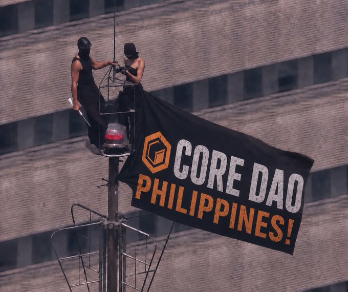 Two masked individuals on a small platform hoist a large black flag reading "CORE DAO PHILIPPINES!" against a tall building backdrop, visually launching the campaign banner referenced in the post inviting builders, creators, and believers to join the Core DAO Philippines community.