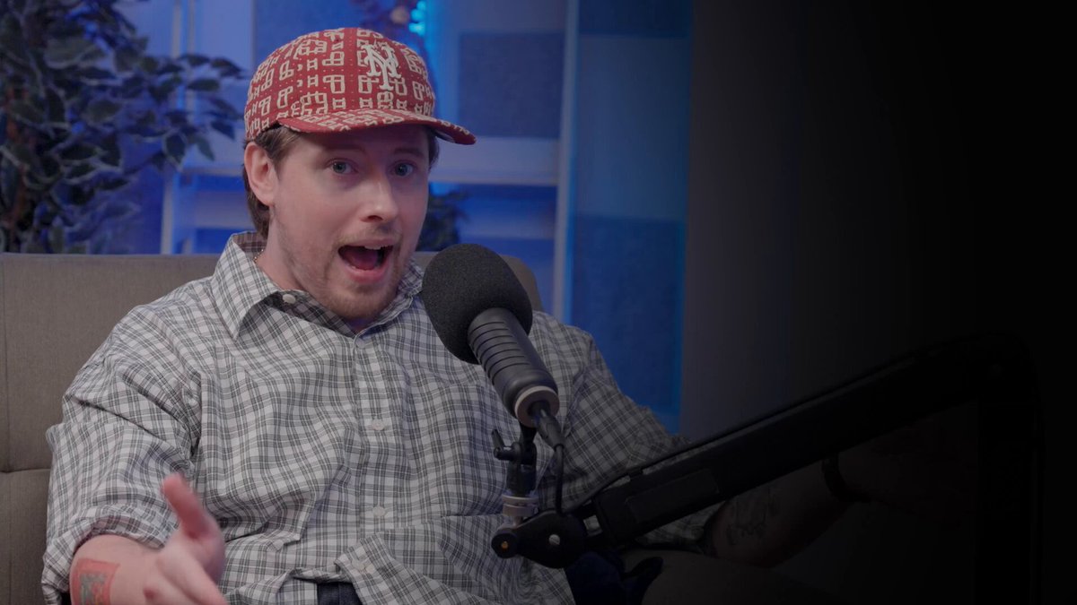 The image features a man wearing a red hat, sitting down with his mouth open as if he's speaking into a microphone or recording device. The background of the photo appears to be a dark room, which could potentially be an audio studio setting. There is also a potted plant visible in the scene, adding some greenery and life to the environment. The image seems to be related to a podcast post, as indicated by the caption provided.
