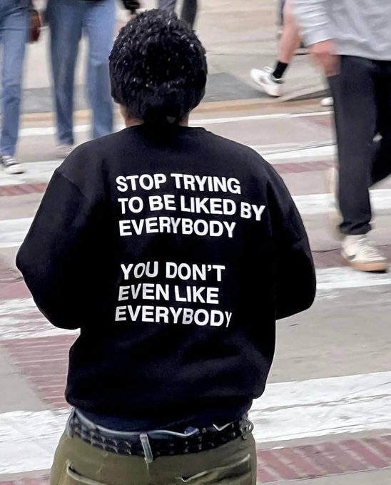 A person walking on a sidewalk wearing a black sweatshirt with white text reading 'STOP TRYING TO BE LIKED BY EVERYBODY / YOU DON'T EVEN LIKE EVERYBODY'.