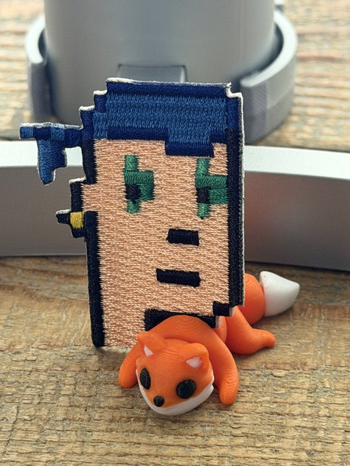 A small 3D-printed pixel-style figure with blue hair and green eyes stands upright on a wooden surface, accompanied by a small orange fox-like creature figurine lying next to it. The scene is set in front of what appears to be a 3D printer.