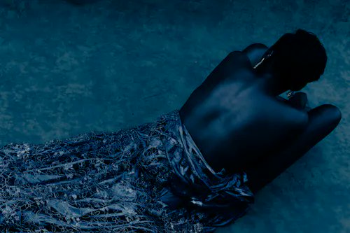 A figure with a bare back sits on the ground at night, partially draped in a textured blue garment, rendered in cool blue tones.