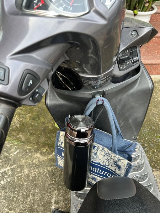 A dark metallic thermos and a blue floral patterned bag hanging from the front hook of a parked scooter or motorcycle.